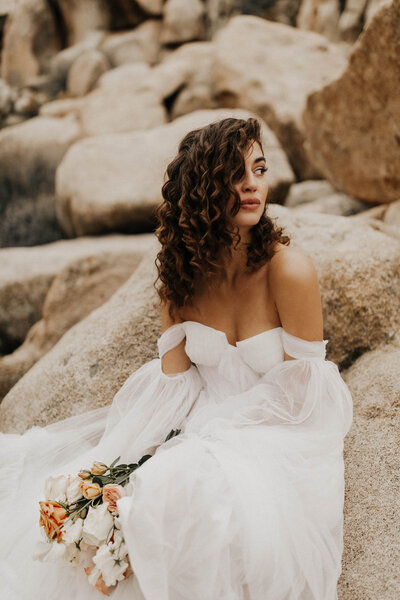 Bride in big white dress holding flowers sits on desert boulders