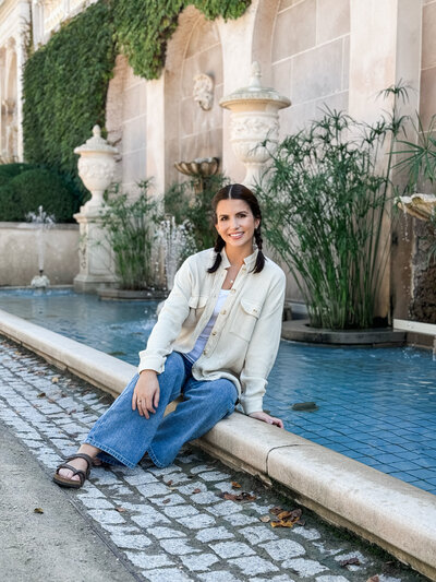 Founder of The Bride's List, Stephanie, sitting by a water feature wearing a tan jacket and denim jeans.