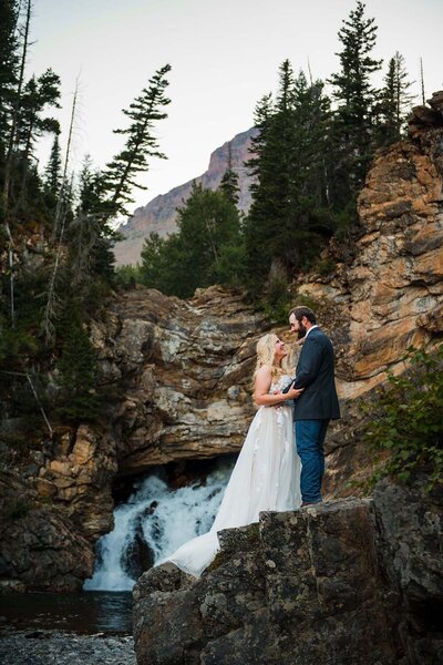 A couple smiles at on another above a scenic waterfall in Glacier National Park in Montana.