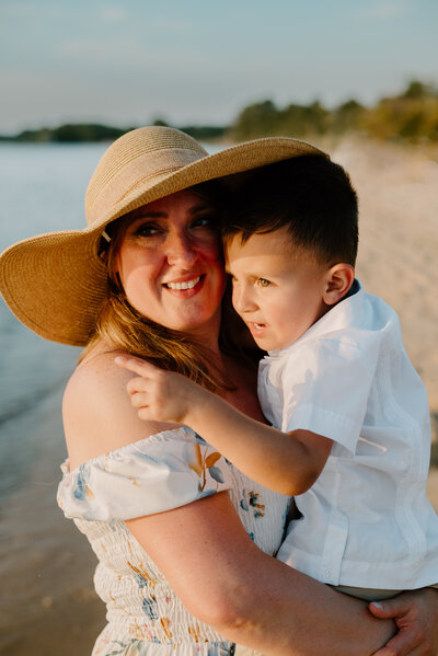 Mom holding toddler son next to the ocean on the beach with the sun shining on them while they are wearing white.