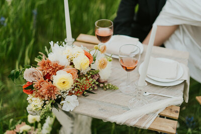 Wedding day detail shot. A close up of a table that has a lacy tablecloth over it. On top is a bouquet of flowers, dishware, and a few glasses of wine.