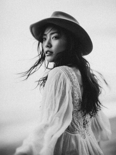 Black and white photo of a woman with long hair in a wide-brimmed hat and lace-trimmed dress, looking over her shoulder—capturing the timeless style that inspires Sweet Pea Ink Creative’s brand design.