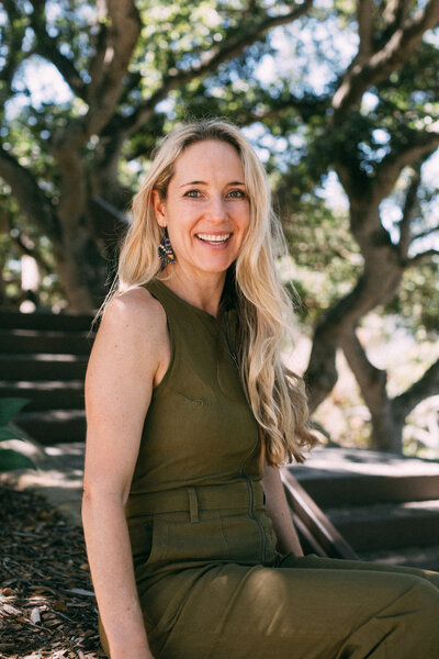 Dr. Diana Hill, ACT Psychologist Leadership Coach, speaking in a red dress with a blue curtain in the background in a green jumper sitting in front of oak trees