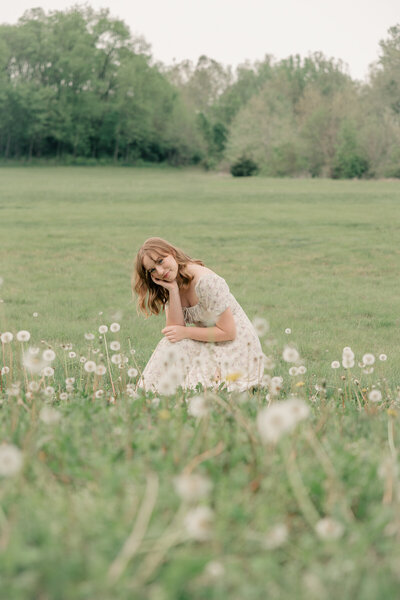 young woman in a cream floral dress crouches in a field of dandelions, Indianapolis senior photography