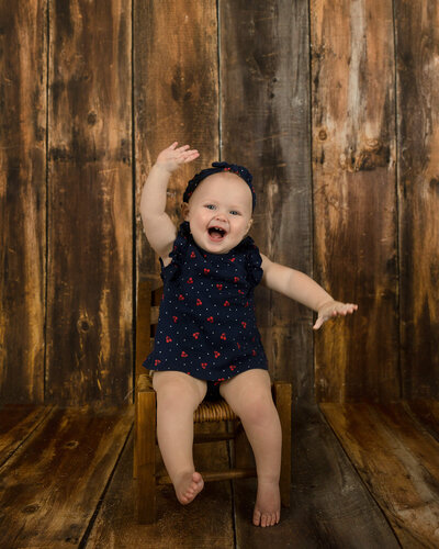 1 year old happy girl smiling and waving as she sits on a little brown chair with wood background.