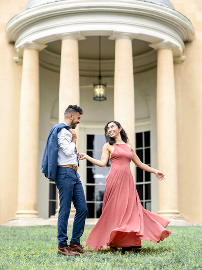 A couple dances joyfully on grass in front of a neoclassical building with tall columns. The woman is wearing a flowing coral dress, and the man is in a white shirt and blue jacket. Both are smiling and holding hands.