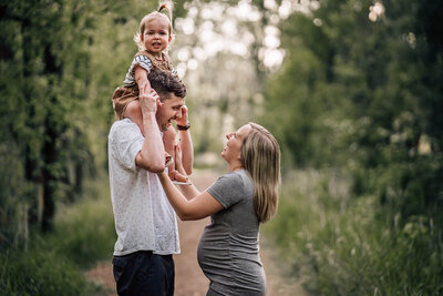 An expecting couple stands facing each other and laughing, with the toddler daughter sitting on Dad's shoulders during a photoshoot at Roberts Bird Sanctuary in Minneapolis.