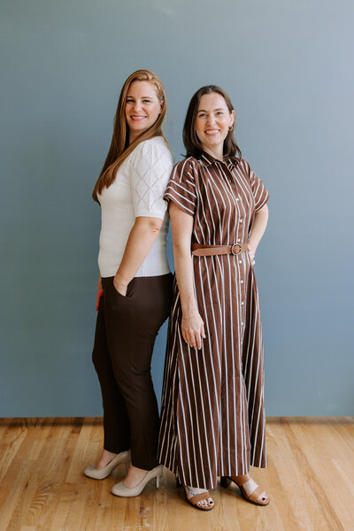 Wedding planners Bridget And Alicia stand back to back while posing in front of a gray-blue wall.
