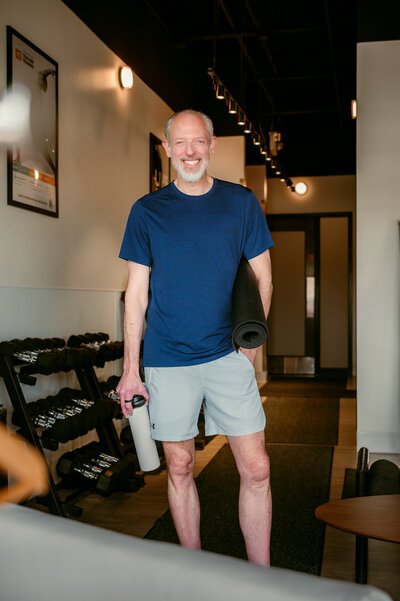 After online dating photo of man smiling confidently in gym setting, holding yoga mat and water bottle in athletic attire
