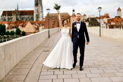 Ein Brautpaar spaziert voller Freude Hand in Hand über die Steinerne Brücke in Regensburg. Die Braut wirft den Arm mit dem Brautstrauß jubelnd nach oben.