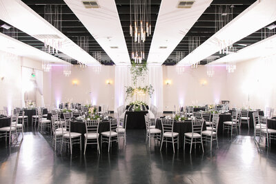 An elegant reception space at SoHo63, a Chandler wedding and event venue, featuring round tables with black linens, silver chiavari chairs, and a dramatic central head table topped with greenery and white draping. Modern chandeliers and soft uplighting complete the stylish, romantic look.