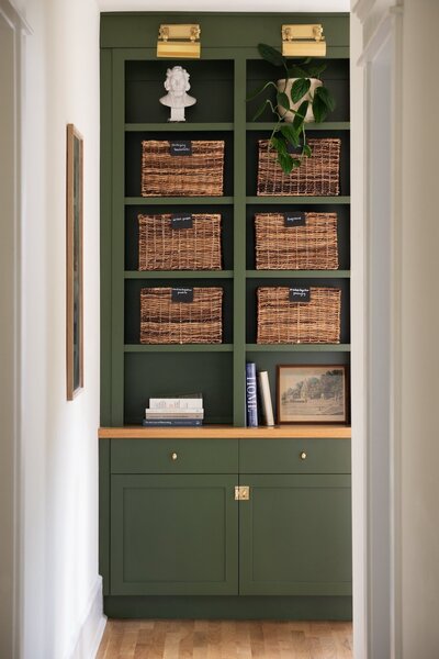 Close-up of green-painted built-in bookcase with wicker basket storage, statement books, and framed decor in upstairs hallway nook.