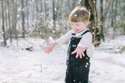 Joyful childhood moment captured in the snow—this outdoor winter portrait features a young boy in classic overalls, laughing as snowflakes fall around him. Birmingham, AL family photographer capturing natural, light-filled moments of childhood wonder.