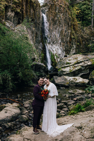 Two brides stand cheek to cheek holding a bouquet with Drift Creek Falls flowing behind them after saying their vows at their Oregon Coast hiking elopement. 