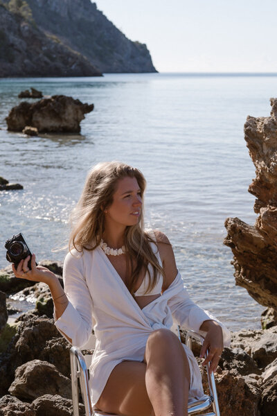 An Ibiza photographer is sitting on a chair on the rocks with a camera in hand.