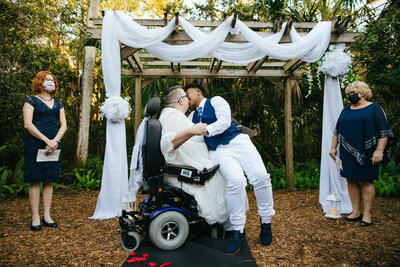 Two Brides kissing during their intimate wedding ceremony. One Bride is in a wheelchair and the other Bride is sitting on her lap to kiss her.