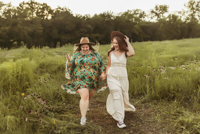 Two sisters in dresses and boho hats hold hands and laugh while walking through a flower field at sunset