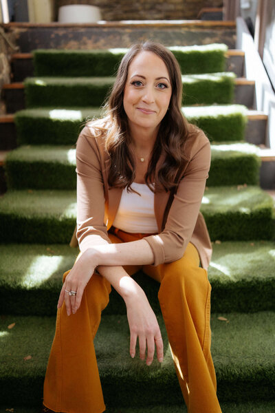A woman with long brown hair sits on green artificial grass steps, wearing a tan blazer, white top, and mustard yellow pants, smiling gently at the camera in natural light. 