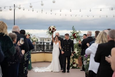 A newlywed couple celebrates their wedding ceremony at the White Room in historic downtown St. Augustine. The bride in a white mermaid-style gown and groom in military dress uniform raise their bouquets triumphantly while walking down the aisle. Wedding guests applaud from both sides under string lights on an outdoor terrace with ocean views in the background.