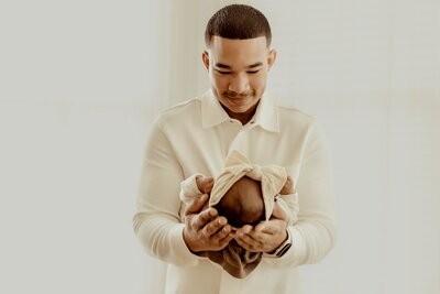 A man in a light-colored shirt gazes at a newborn wrapped in a blanket with a fabric bow on their head, cradled in his hands against a softly lit background.