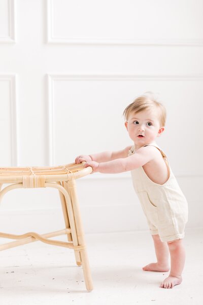 A baby is standing next to a rattan stool in a portrait taken by Charlotte photographer.