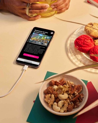 A smartphone displaying the homepage of the B Town Social website is placed on a table next to a bowl of mixed nuts and a plate of colourful cookies. A person's hands holding a glass cup are visible in the background. The phone screen shows an image of a vibrant street scene and a welcome message for the food blog, emphasising Birmingham's local food and drink scene.