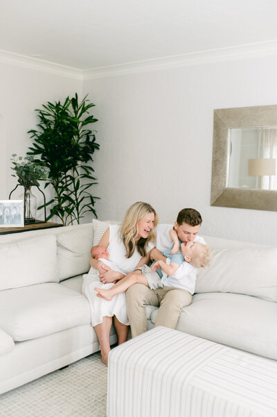 Pulled back shot of a mom and dad on a couch. Mom is holding baby and the dad is holding his toddler on his lapping, dipping him back to kiss his cheek and laughing