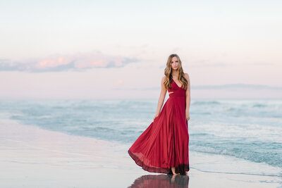 Teen girl in flowing red dress standing barefoot at water’s edge during golden hour senior pictures at Folly Beach near Charleston

