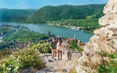 A couple hiking up a rocky trail overlooking a wide river valley with green hills, vineyards, and a small village below.