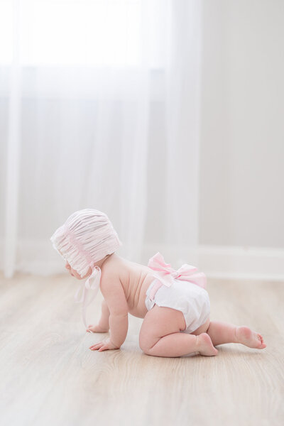 Baby girl crawling on the floor with a pink bow diaper cover and pink bonnet for her milestone session
