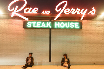 A couple poses in front of the neon-lit Rae and Jerry’s Steakhouse sign, evoking a nostalgic romance.