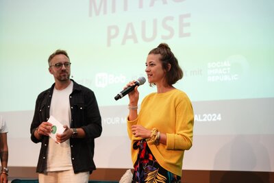 Eventmoderatorin Hanna Zöller im gelben Pullover bei Co-Moderation auf Bühne, mit Mikrofon in der Hand und Publikum im Hintergrund
