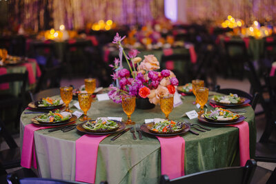 A wedding tablescape with green velvet linens, bright pink napkins, and floral arrangements with bright pinks and oranges