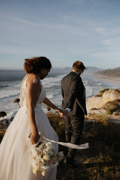 a newlywed couple holding hands and walking along an ocean cliffside 