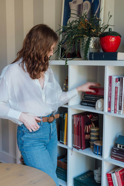 Erin, an interior designer in Portland, in her home reach for a book on shelf with knick knacks. 