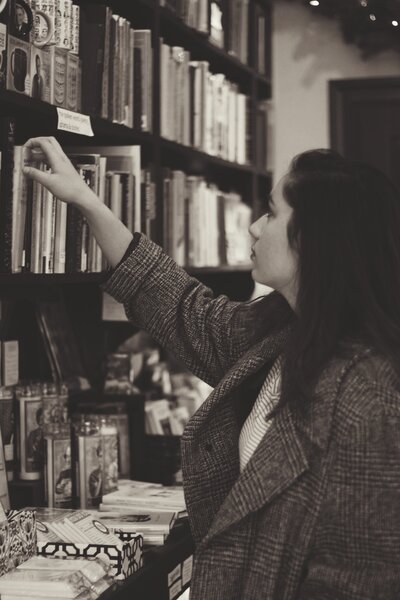 Young woman with oversized tween coat looking at bookshelf reaching to take a book off the shelf