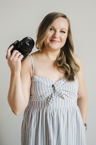Krista Marie, standing in front of a white wall holding her camera near her shoulder, smiling and looking at the camera