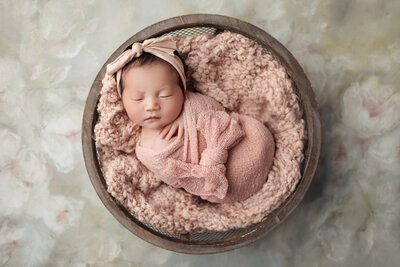 A newborn baby is peacefully sleeping, swaddled in a soft pink wrap and nestled in a wooden bowl lined with a fluffy pink blanket. The baby is wearing a matching pink headband with a bow, adding an extra touch of sweetness to the scene. The bowl is placed on a light, textured background that has subtle floral patterns, creating a gentle and serene atmosphere. The baby's hands are tucked under their chin, and their expression is calm and content, making this a charming and beautifully composed portrait.