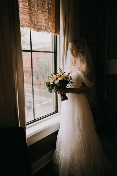 Bride holding her flowers and looking out a window in her wedding dress before her wedding in Pensacola, Florida