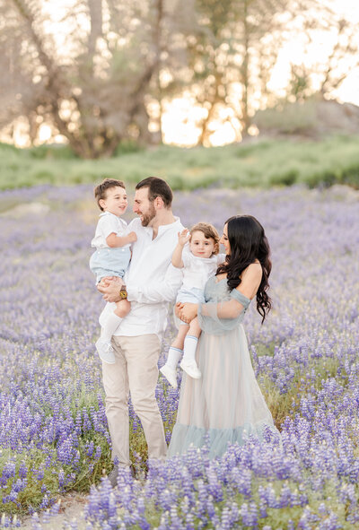 A family of four stand in a field of purple lupines dressed in shades of grey, blue, and white while smiling and holding one another photographed by bay area photographer, Light Livin Photography.