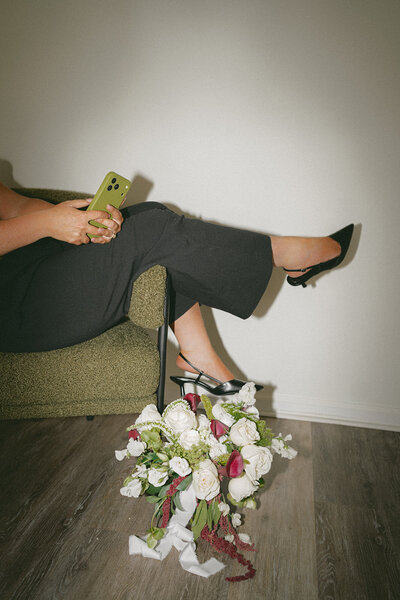 kate of kate's content co sitting on the side of a green chair with her feet in black shoes hanging off the edge while editing client's photos on her green phone after a wedding content creation