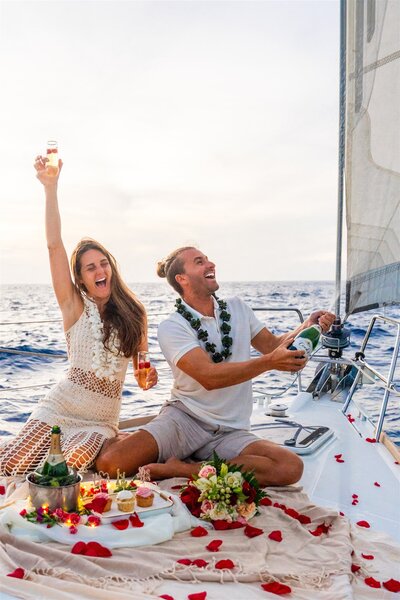 Engaged couple celebrating on a sailboat in Hawaii as the man pops a bottle of champagne and the woman cheers with excitement over the ocean.