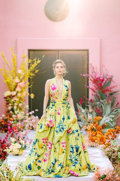 woman standing in green floral dress surrounded by flowers and pink wall
