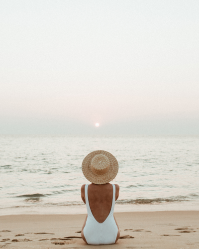 woman in white one-piece swimsuit and sun hat sits on a beach looking out at water