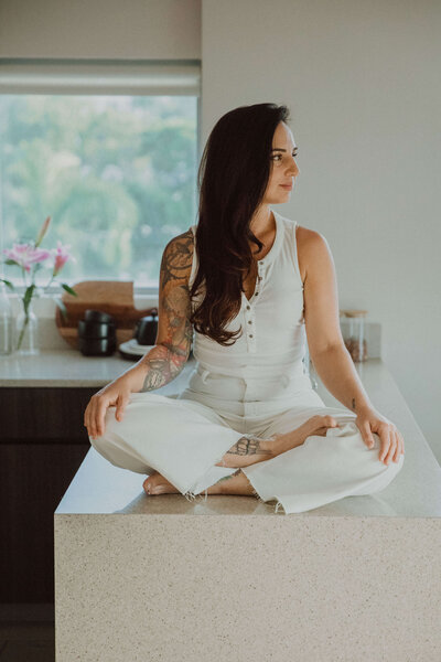 Ayurvedic Skin Specialist, Jen, wearing a white top and pants sitting on a jute rug leaning against a neutral couch