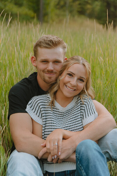 Sitting on the ground in a field of tall grass, a man holds his fiancé between his legs with his arms wrapped around her. 