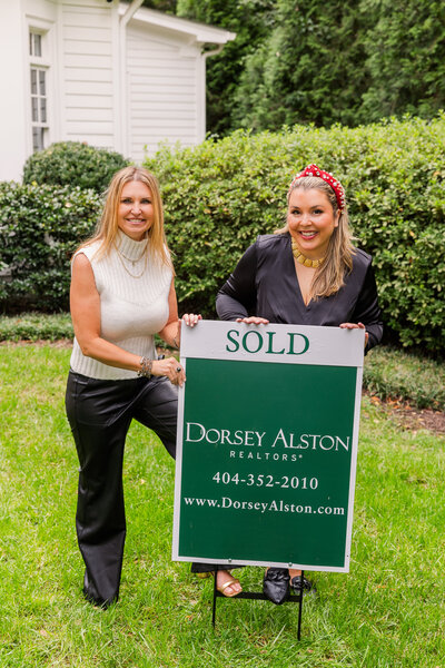 realtor women team wearing coordinated black and white outfits and holding a sold sign in a house front yard during photoshoot
