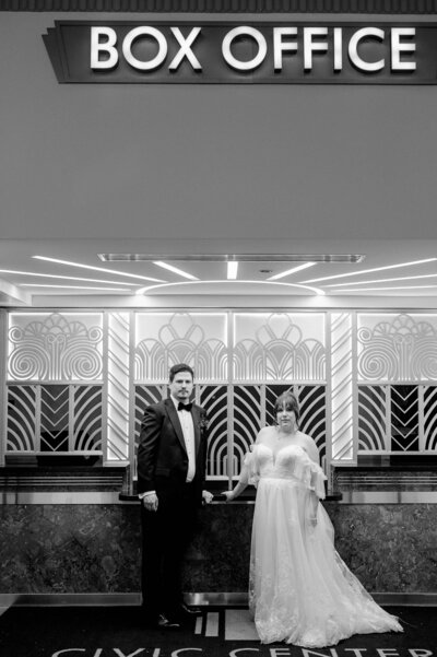 A couple stands in front of an art deco box office at the Civic Center Hall of Mirrors in an editorial wedding portrait.