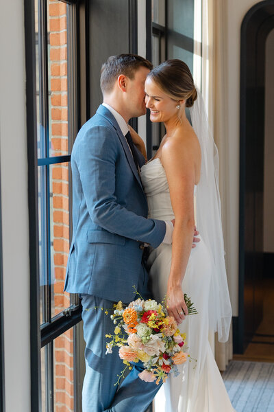 Bride in white dress and groom in blue suit share intimate moment by brick window with colorful bouquet.