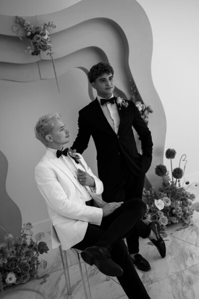 A groom in a white suit sits on a chair, while another groom in a black tuxedo stands beside him, both in a stylish wedding setting.
